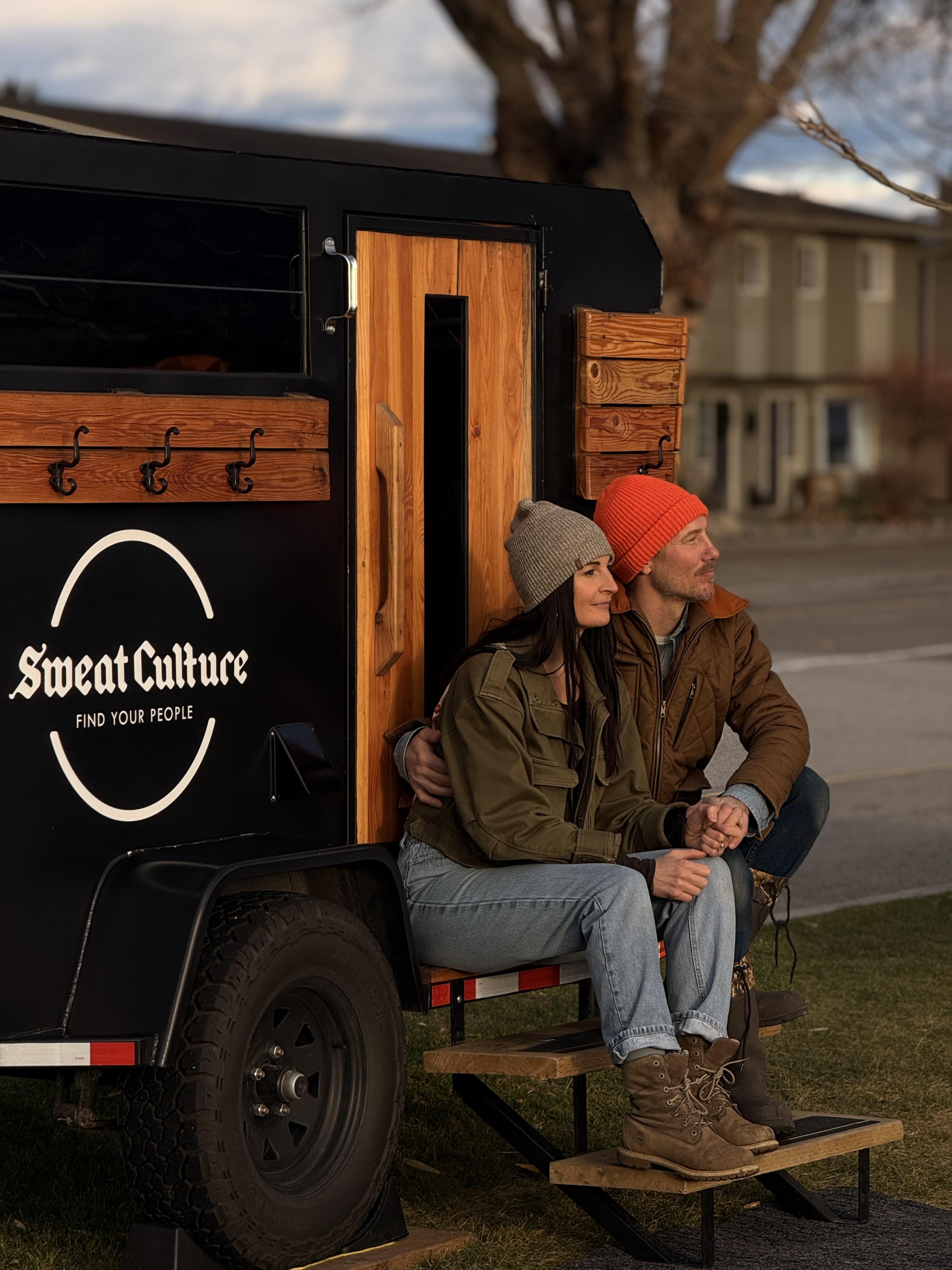 Sweat Culture founders Nina and Braden sitting together at golden hour beside the wood-fired adventure trailer sauna in Peachland, BC