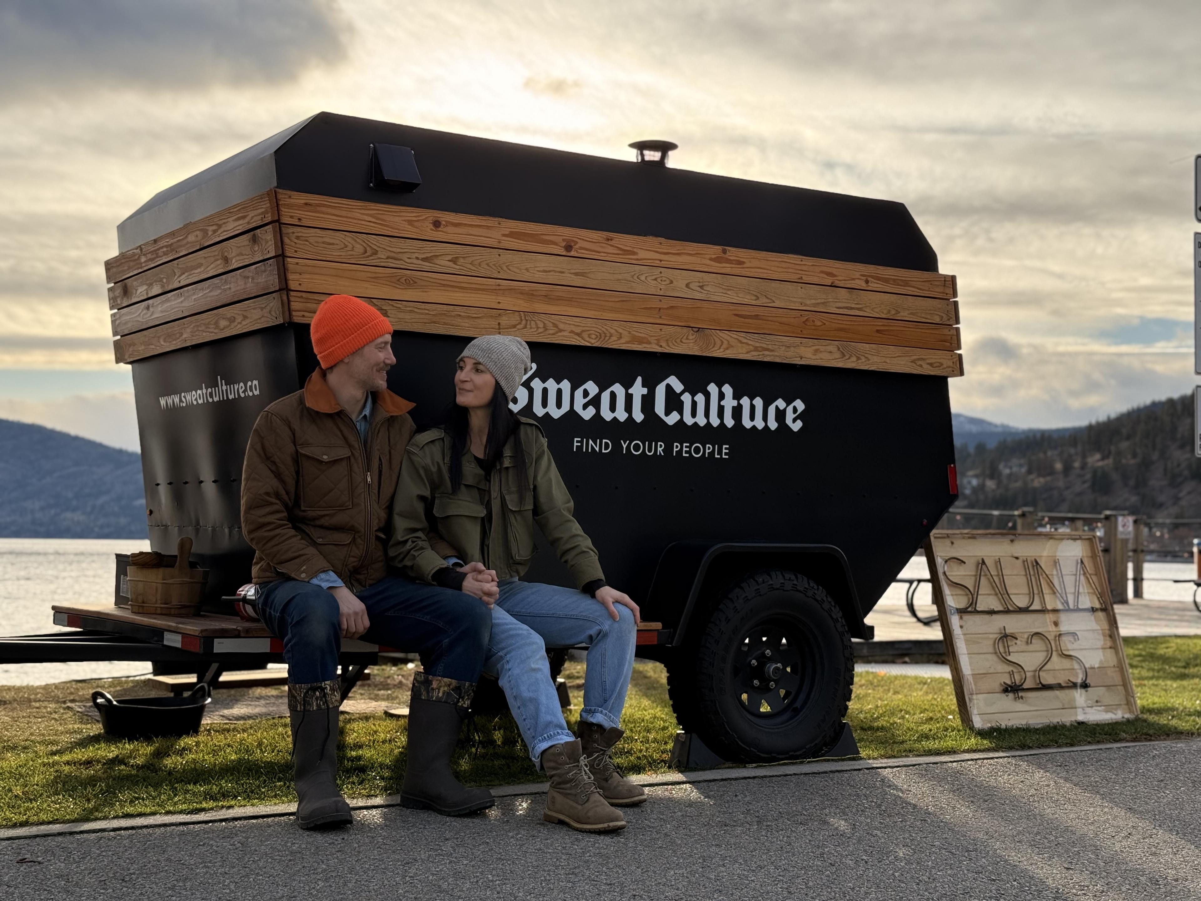 Sweat Culture founders in front of the adventure trailer sauna with Okanagan Lake in the background and a $25 sauna sign in Peachland, BC