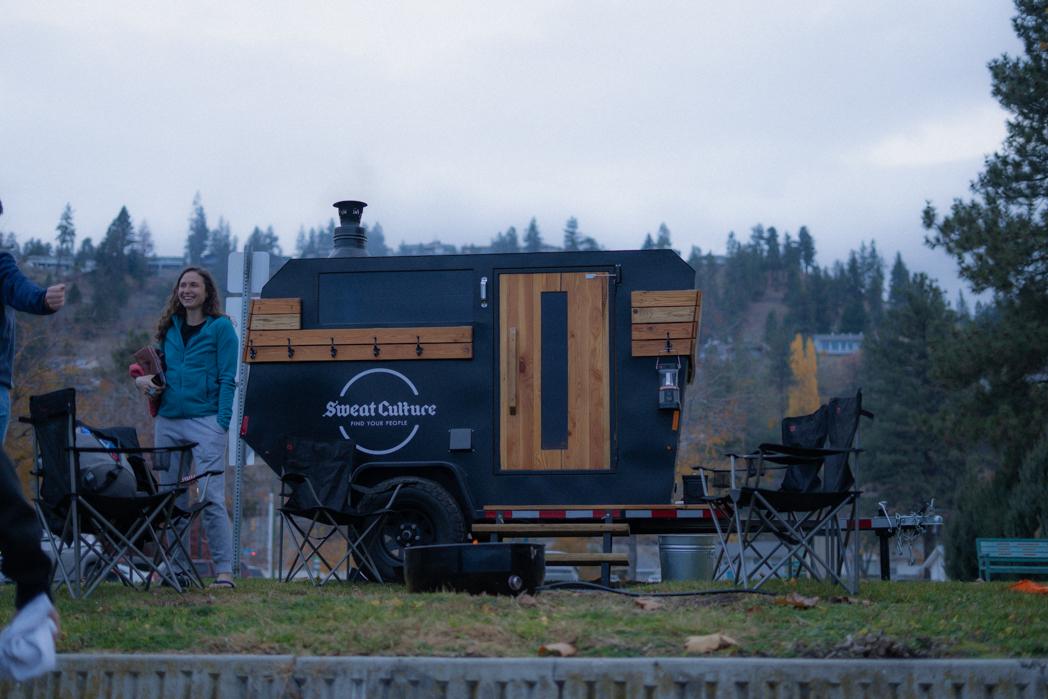 The Sweat Culture wood-fired sauna trailer set up on Beach Ave in Peachland at dusk with autumn trees and Okanagan mountains in the background