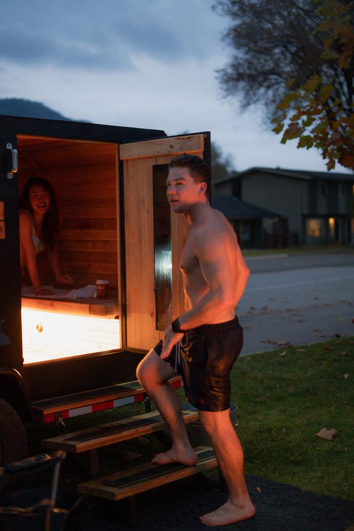 Guest stepping out of the Sweat Culture sauna at night, fire pit burning in the foreground on Peachland Beach, Okanagan Lake