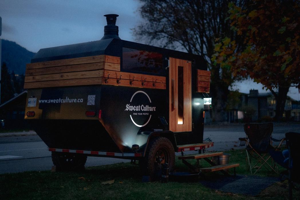 The Sweat Culture wood-fired adventure trailer sauna glowing at night on Beach Ave in Peachland, BC with Okanagan Lake behind it
