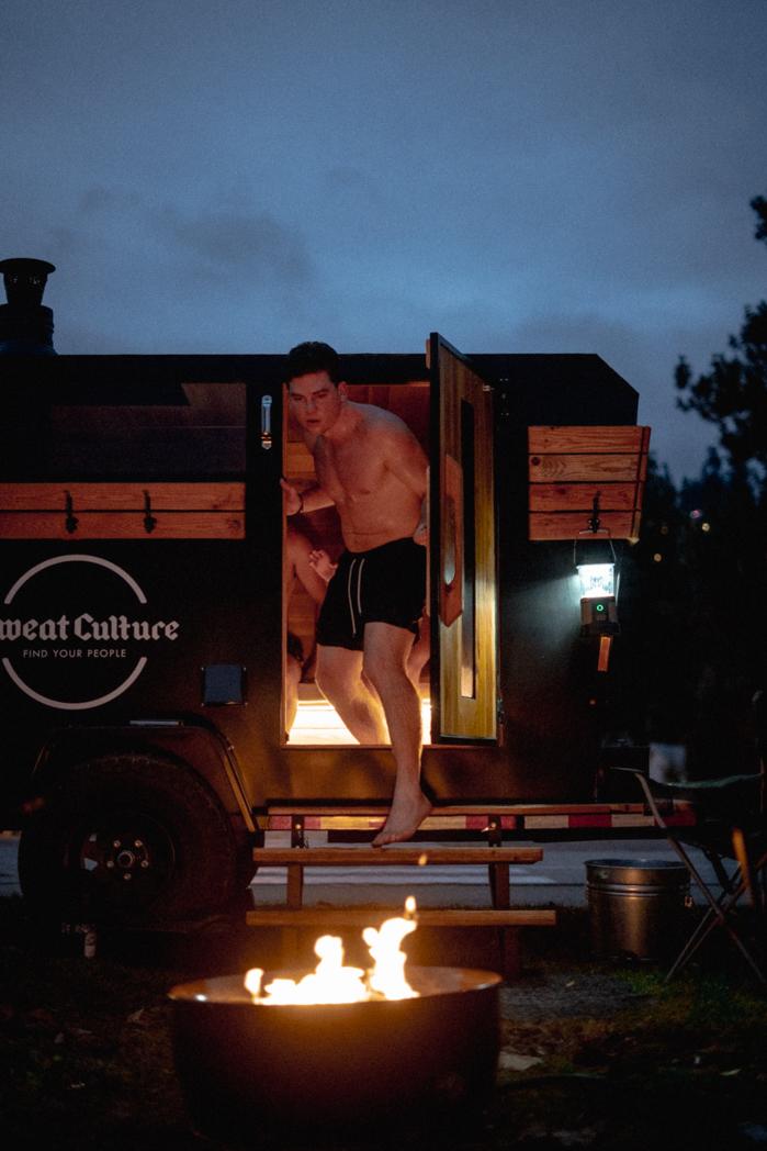 Guest stepping out of the glowing Sweat Culture sauna door at night, fire pit lit in the foreground, outdoor sauna session in Peachland, BC
