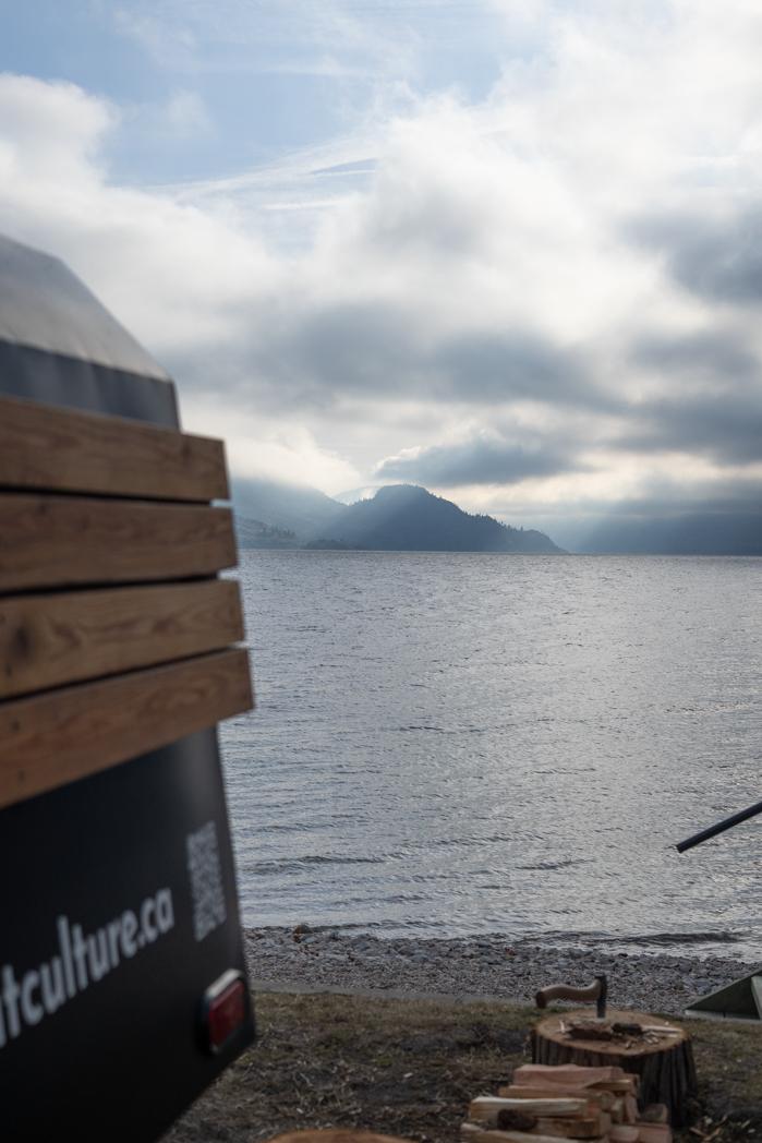View of Okanagan Lake and the mountains from beside the Sweat Culture adventure trailer sauna on the shore in Peachland, BC, with firewood stacked nearby