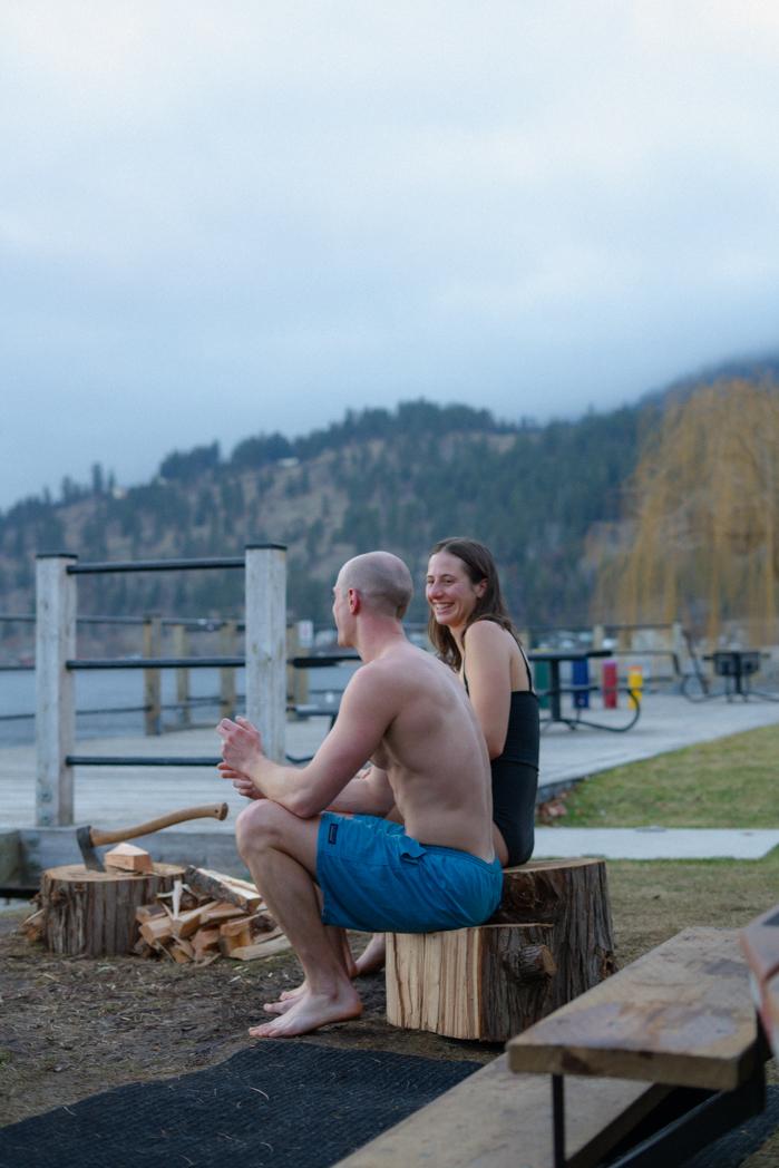 Couple laughing and relaxing on wooden stumps by Okanagan Lake after a Sweat Culture sauna and cold plunge session in Peachland, BC