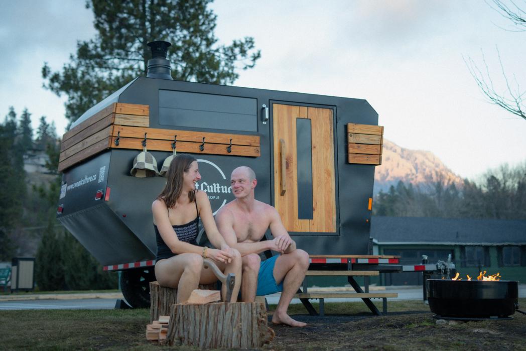 Couple sitting in front of the Sweat Culture adventure trailer sauna with the fire pit burning and snow-capped Okanagan mountains at sunset