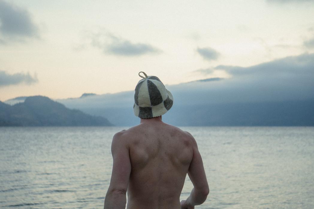 Guest wearing a traditional Finnish sauna hat facing Okanagan Lake at dusk, preparing for a cold plunge after a Sweat Culture sauna session in Peachland