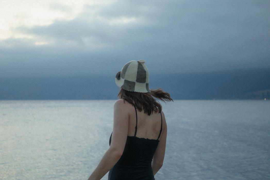 Guest in a sauna hat gazing out over Okanagan Lake at dusk from Peachland Beach, cold plunge therapy with Sweat Culture