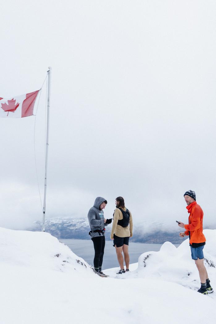 Sweat Culture run club members standing beside a Canadian flag on a snow-covered summit overlooking Okanagan Lake, winter run club in Peachland, BC